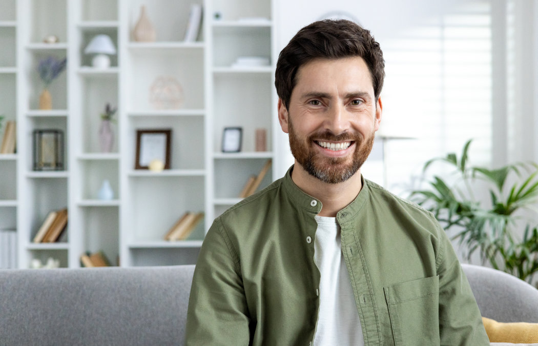 A man with brown hair and a beard, wearing a green shirt over a white t-shirt, sits on a gray couch in a modern living room with shelves in the background., Indian Land, SC
