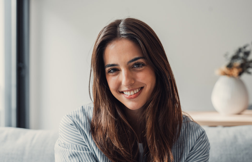 A young woman with long brown hair sits indoors, smiling at the camera. There is a white vase with dried flowers in the blurred background., Indian Land, SC