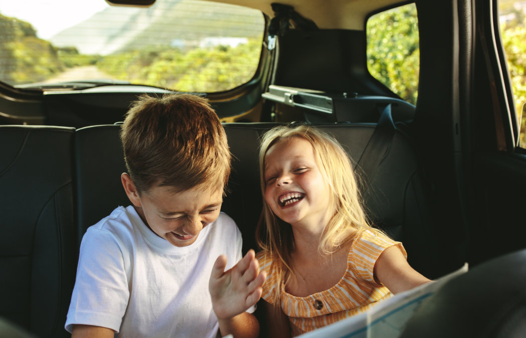 Two children sit in the backseat of a car, laughing and smiling, with greenery visible through the rear window., Indian Land, SC