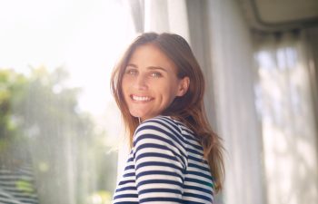 Woman, smile and portrait in house for relax and calm for holiday and cheerful on blurred background. Young person and happy with window for view of garden or backyard with sunlight in lounge